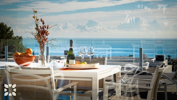 A dining table with a bowl of oranges, a vase with dried flowers, a bottle of wine, and two wine glasses on a terrace with a view of the ocean. - Villa YANSA by Abahana Villas (Benitachell)