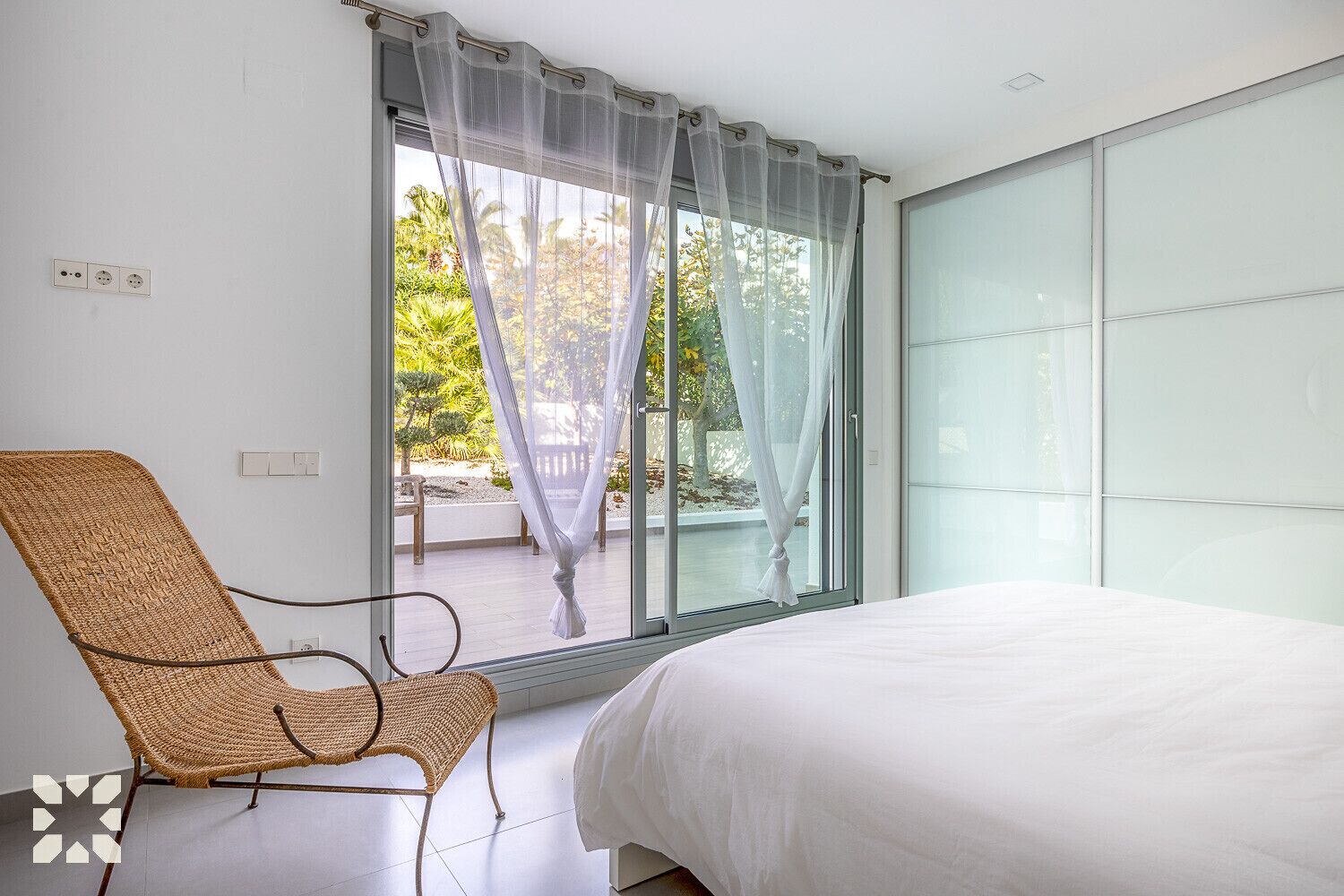 Een slaapkamer met een bed, een rieten stoel, een groot raam met dunne gordijnen en uitzicht op een tuin.