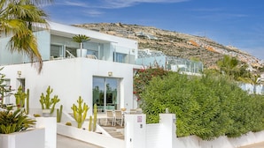 A modern white building with a glass facade, a patio with chairs, and a variety of cacti and shrubs. - Villa YANSA by Abahana Villas (Benitachell)