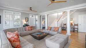 Ein geräumiges Wohnzimmer mit einer großen Eckcouch, einem Couchtisch und zwei Zierteilen. Im Hintergrund sind zwei Deckenventilatoren, eine Treppe und ein Essbereich zu sehen.