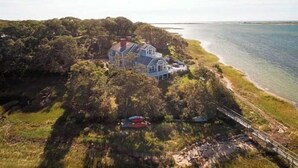 A large, two-story house with a prominent balcony, situated on a cliff overlooking a body of water.