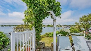 A view of a balcony with a white gate, a pergola covered in greenery, and a row of sun loungers overlooking a body of water with boats.