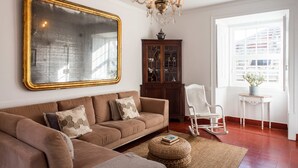 A living room with a large mirror, a chandelier, a wooden cabinet, a rocking chair, and a window with a view of buildings.
