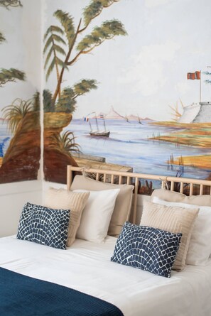 Ein Schlafzimmer mit Wandgemälde einer Strandansicht, einem Bett mit weißer Bettwäsche und gemusterten Kissen sowie einem hölzernen Kopfteil.