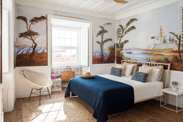 A bedroom with a bed, a chair, a nightstand, a window, and a mural on the wall.