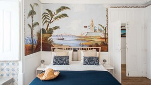 A bedroom with a mural of a coastal scene, a bed with a blue bedspread, and a straw hat on the bed.