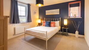 A bedroom with a bed, bedside lamps, a window with curtains, a radiator, a wall-mounted shelf with a plant, and a painting. - Host Stay The Sanctuary (Whitby)