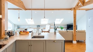 Cozinha moderna com ilha central, vigas de madeira, claraboias e área de jantar com sofá e televisão.