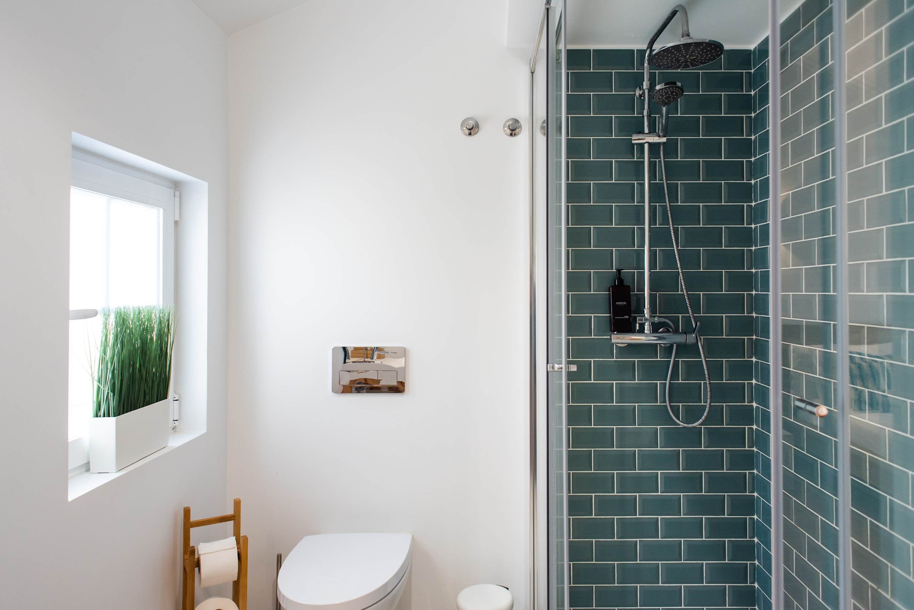 A bathroom with a white toilet, a wooden chair, a window with a plant, and a shower with blue tiles.