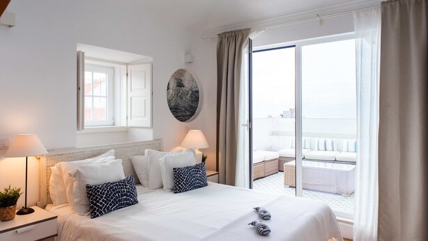 Quarto de hotel com uma cama grande, duas luminárias de cabeceira, vista para a varanda e um quadro redondo na parede.