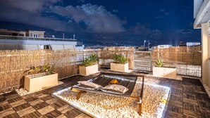 Eine Dachterrasse mit Liege, Topfpflanzen und Blick auf die beleuchtete Stadt.