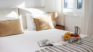 Ein ordentlich bezogenes Bett mit weißen und beigen Kissen, ein Nachttisch aus Holz mit Kaffeemaschine und Croissants sowie eine Zeitschrift auf dem Bett.