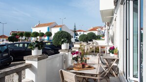 Ein Balkon mit Tisch und Stühlen, Blumentöpfen und Blick auf die Straße mit parkenden Autos und Gebäuden.