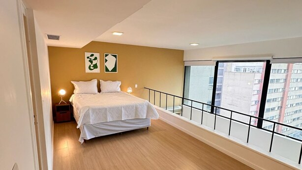 Une chambre à coucher avec un lit, une table de chevet, une lampe et une vue sur les bâtiments, visible à travers la balustrade du balcon.