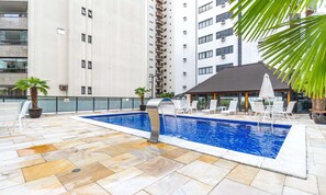 A swimming pool with a water feature, surrounded by lounge chairs and umbrellas, with tall buildings in the background. - Victoria Charm Stylish 1 BDR in Itaim Bibi (São Paulo)