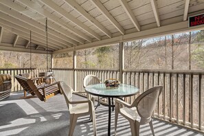 A covered outdoor seating area with a table and chairs, a hanging swing, and a view of trees through the railing. - New Listing! Hot Tub, Foosball, Fireplace, Pool Table, & Free Attractions! (Sevierville)