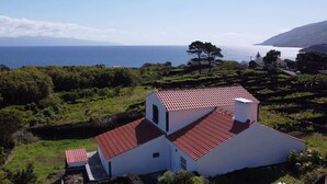 A white building with a red roof, surrounded by greenery and overlooking a body of water. - Liiiving in A Ores - Ocean View House (Sao Roque do Pico)