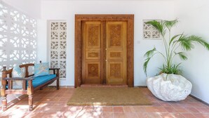 A wooden door with intricate carvings, a bench with cushions, a large potted plant, and a decorative wall panel. - Lazy Days (Diani Beach)