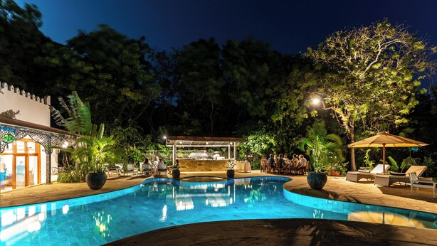 A swimming pool with lounge chairs and a covered seating area, surrounded by trees and a building with a decorative facade. - Lazy Days (Diani Beach)