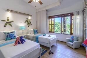 A room with two beds, a small table, a chair, and a window with a view of greenery. - Lazy Days (Diani Beach)