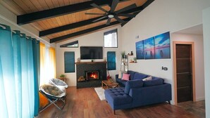 A living room with a fireplace, a blue sofa, and a ceiling fan. - Beautiful Renovated Cozy Forest Home in Crestline with Hot Tub (Crestline)