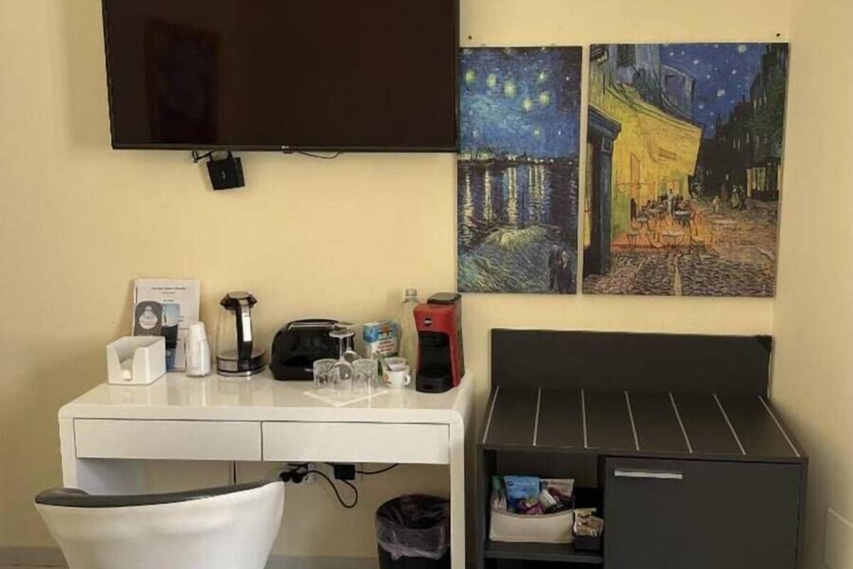 A room with a flat-screen TV mounted on the wall, a white desk with a coffee maker, glasses, and a trash bin. There are two paintings of starry night scenes on the wall.
