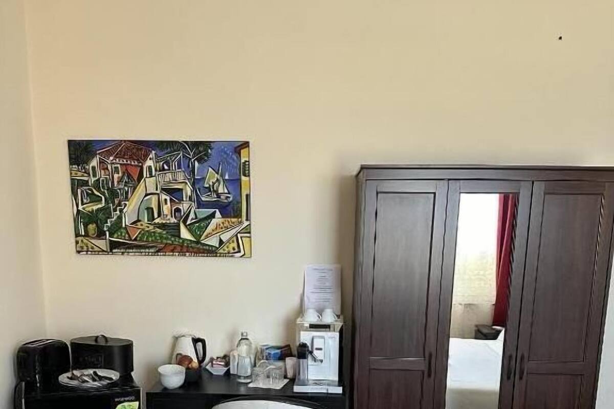A hotel room with a bed, a desk with a coffee maker, a chair, a wardrobe, and a painting on the wall.