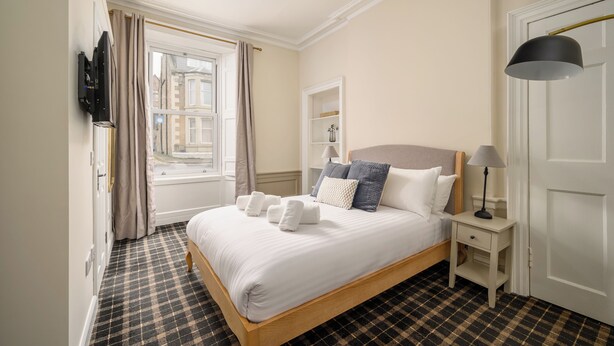 Ein ordentlich eingerichtetes Schlafzimmer mit kariertem Boden, einem Bett mit weißen Leinen und mehreren Kissen, einem Nachttisch mit Lampe und einem Fenster mit Vorhängen.