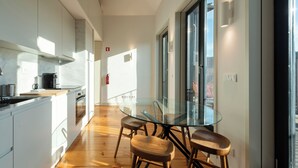 A modern kitchen with white cabinetry, a glass dining table, wooden stools, and a large window providing natural light. - Unusual House (Porto)
