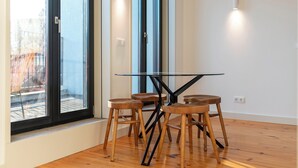 A modern room with a glass table and wooden stools, large windows with blinds, and a white wall with a light switch. - Unusual House (Porto)