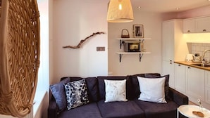 Un salon moderne avec un canapé foncé, des coussins décoratifs, un luminaire suspendu et un coin cuisine doté de meubles blancs et d’un plan de travail.