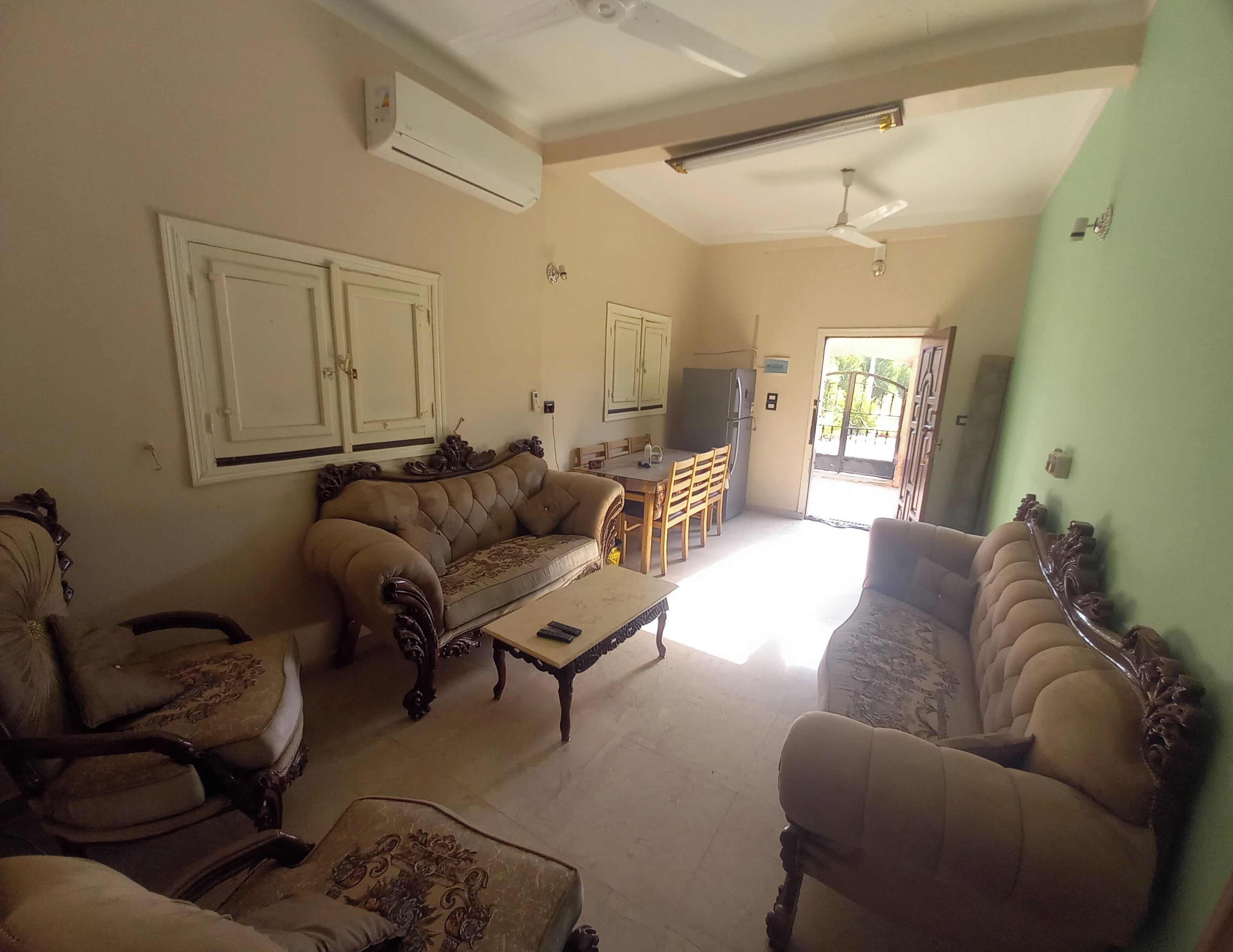 Una sala de estar con un sofá, sillones, una mesa de centro y un televisor. Hay un comedor con una mesa y sillas. La habitación cuenta con un equipo de aire acondicionado y un ventilador de techo.