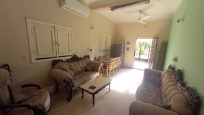 A living room with a sofa, armchairs, a coffee table, and a television. There is a dining area with a table and chairs. The room has an air conditioning unit and a ceiling fan.