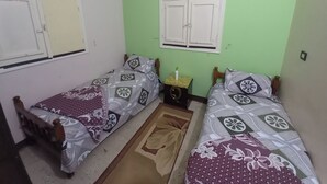 A small, clean room with two single beds, a window with white shutters, and an air conditioner on the wall.