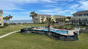 A pool area with lounge chairs and palm trees, surrounded by apartment buildings with a view of the ocean.