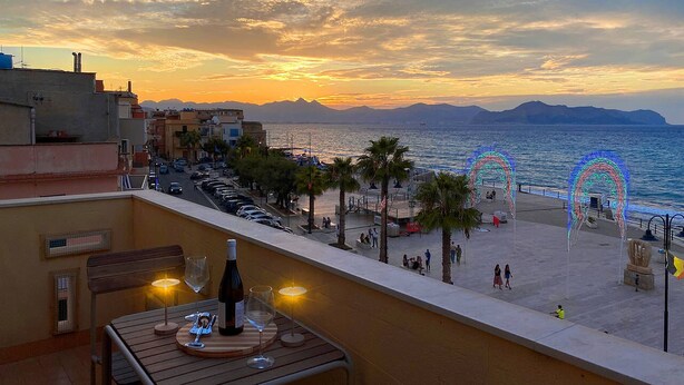 A balcony with a table, a bottle, and a glass, overlooking a beach with palm trees and a sunset. - Solemar Sicilia - Casa Carina (Bagheria)