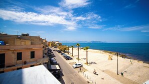 A coastal street with parked cars, a pedestrian walkway, and a view of the sea and distant mountains.