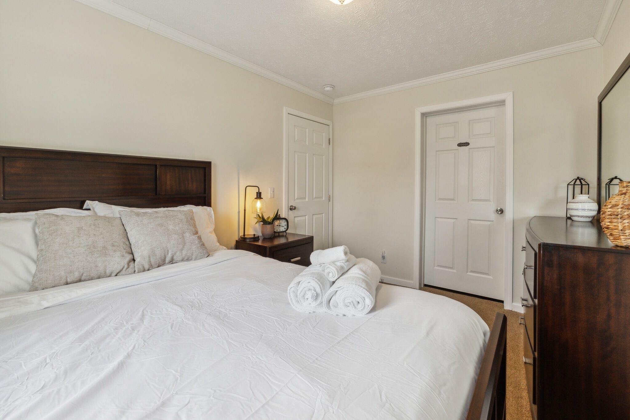 A neatly made bed with white linens and pillows, a bedside table with a lamp and a clock, and a dresser with a mirror and a basket.