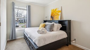Un dormitorio con cama, ventana con cortinas, un cuadro en la pared y una mesita de noche.