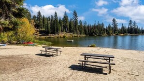 Une plage de sable avec deux tables de pique-nique et un lac paisible, entouré d’une forêt dense.