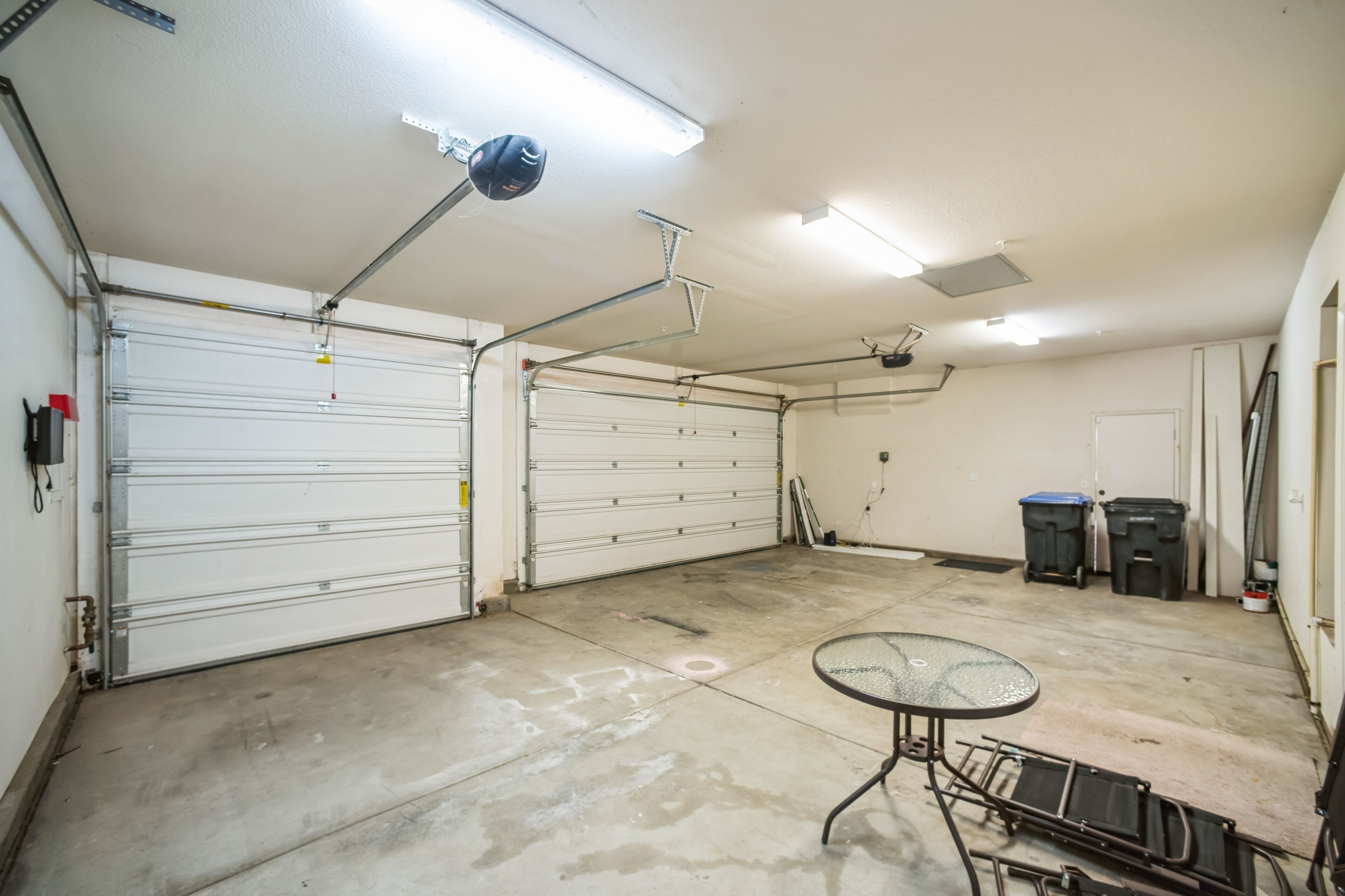 A spacious garage with two large white garage doors, a concrete floor, and a small table with chairs.
