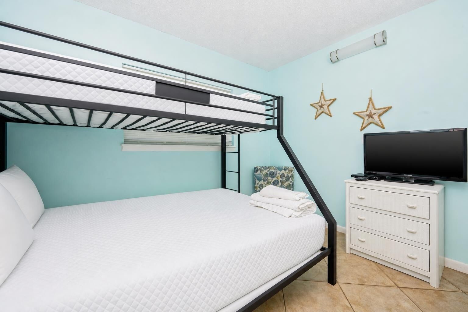 A bunk bed with a flat bed below, a white dresser with drawers, a television on top, and two star decorations on the wall.