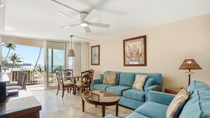 A spacious living area with a blue sofa, a glass coffee table, and a ceiling fan. - Kamaole Nalu, #304 2 Bedroom Condo (Kihei)