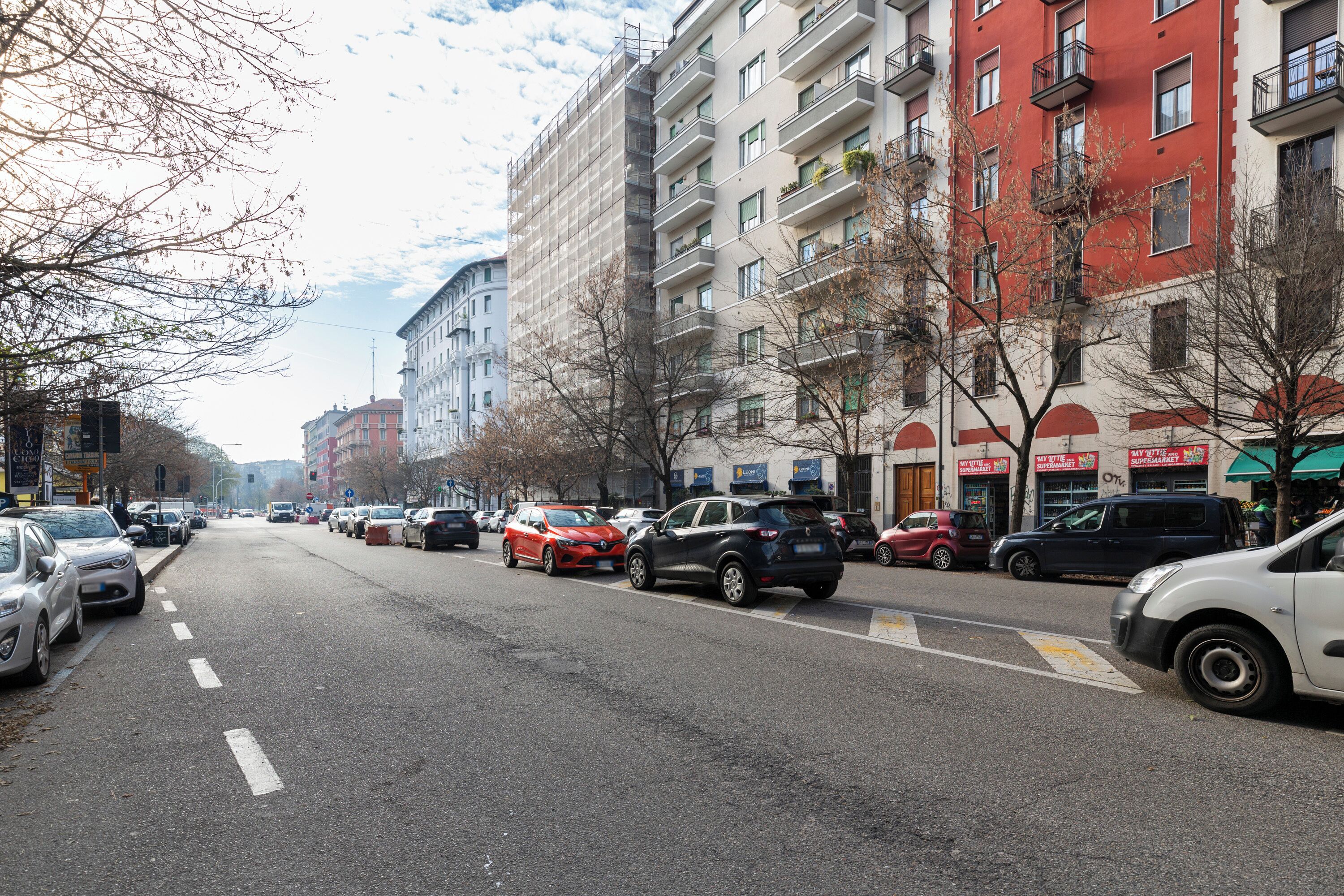 Une rue bordée d’immeubles à plusieurs étages, de voitures garées et d’arbres sans feuilles.