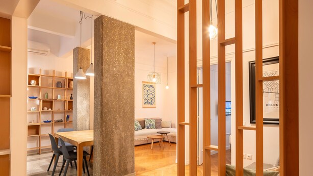 A modern living room with a wooden dining table, chairs, and a sofa. There is a bookshelf, a hanging light fixture, and a wooden partition. - Historic 2BR Oasis in Kolonaki (Athina)