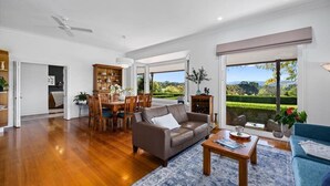 A spacious living room with a wooden floor, a brown leather sofa, a blue armchair, and a dining area with wooden furniture. - Hubbard Hill Yarra Glen Oasis: 4-Bdrm Home with Pool & Stunning Valley Views (yarra glen)