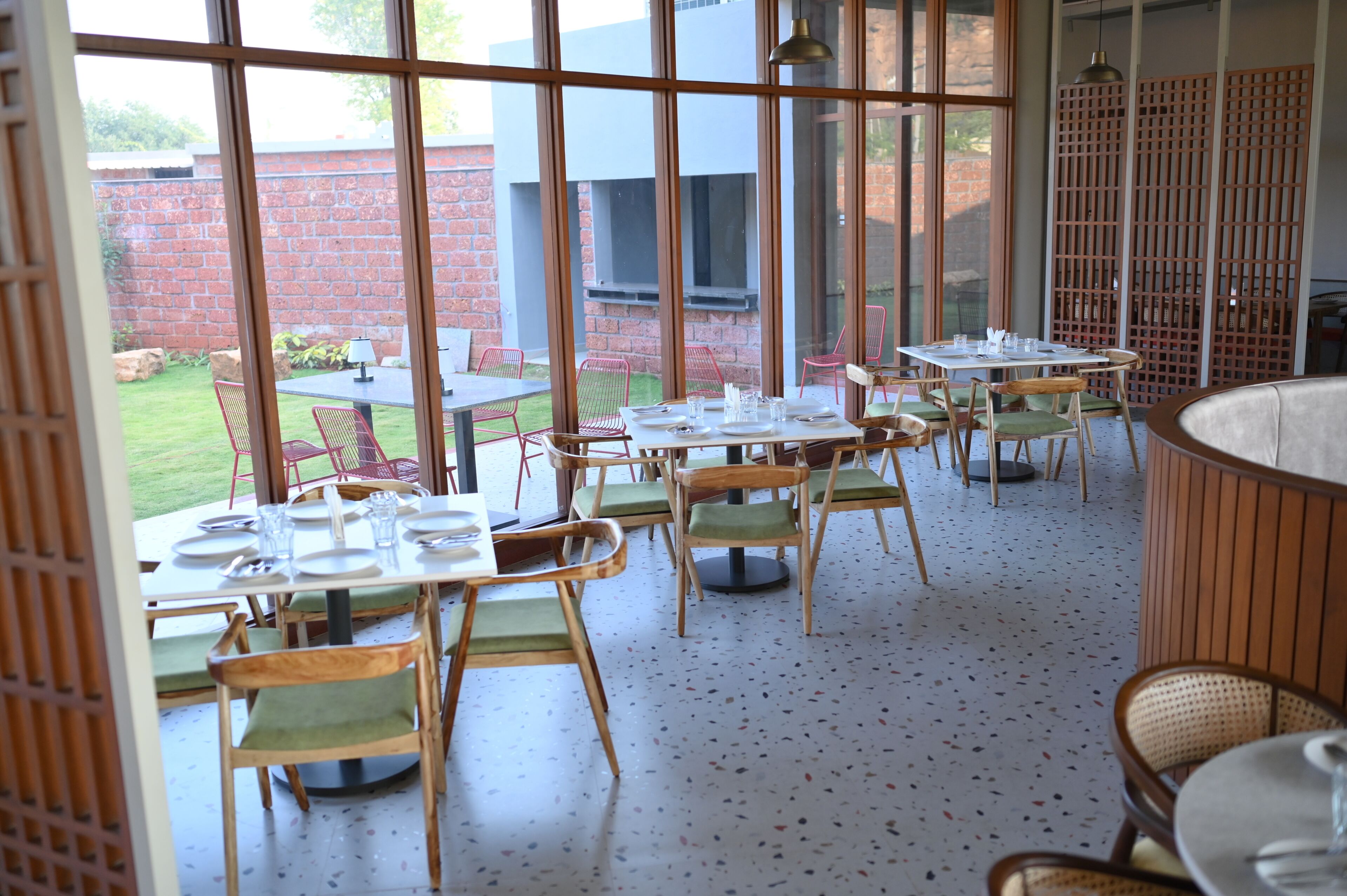 Un comedor interior con mesas y sillas, amplias ventanas y vistas a una pared de ladrillo y vegetación exterior.