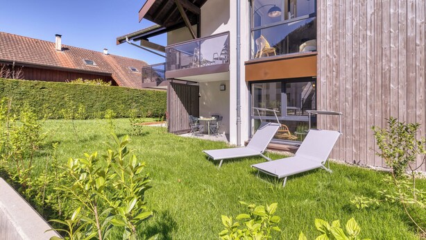 Una casa moderna de dos pisos con un balcón, un césped cuidado y dos sillones de descanso.