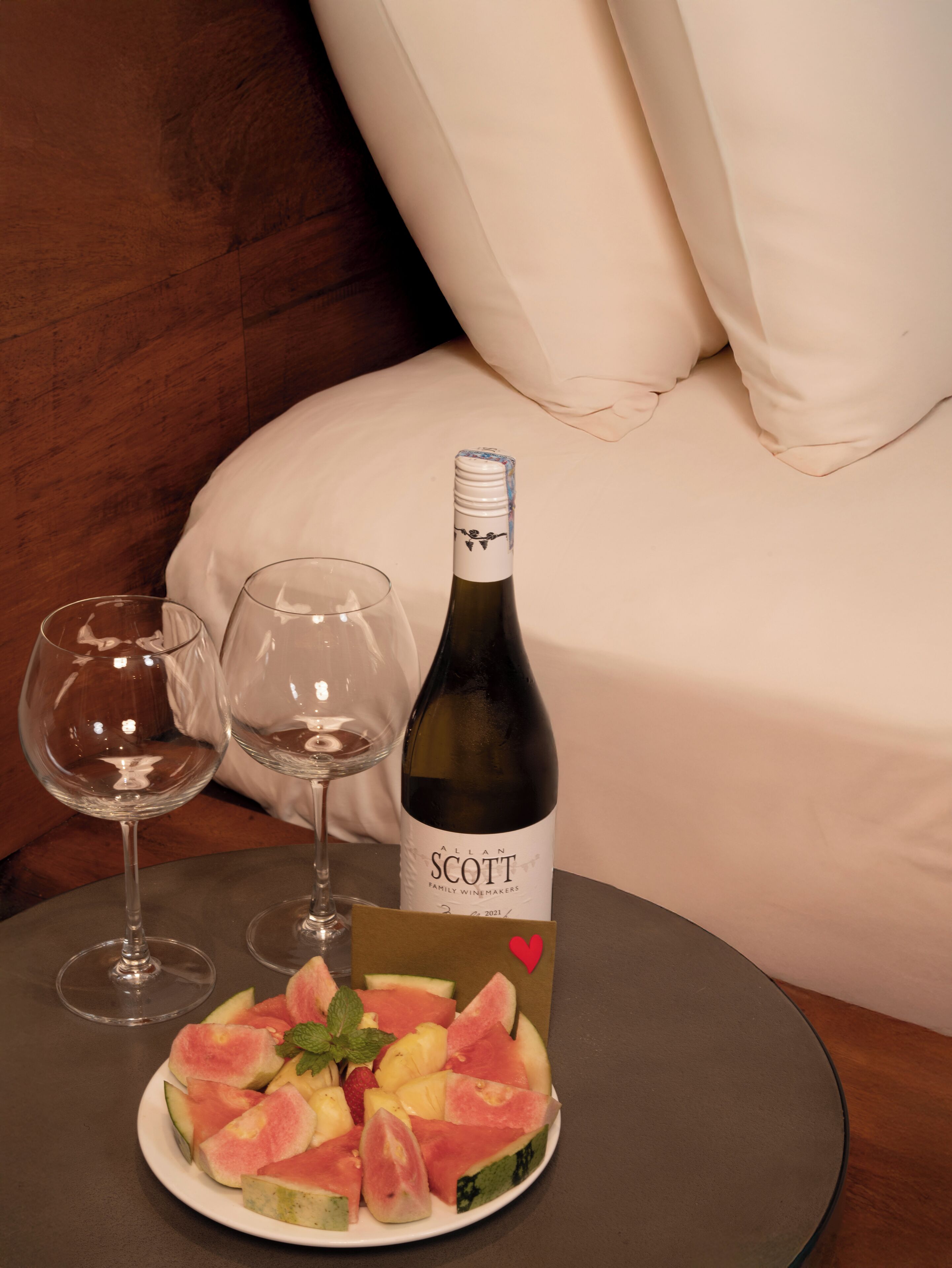 A bottle of wine, two glasses, and a plate of fruit on a table in a room with a bed and white curtains.