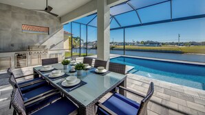 Un espace repas extérieur moderne, doté d’une table et de chaises en verre, d’un barbecue et offrant une vue sur une piscine et un jardin paysager.